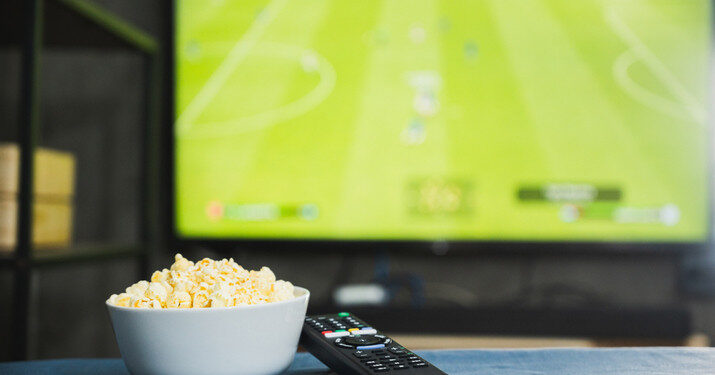 Popcorn and television remote control on football program tv screen background. Watching tv relax concept.
