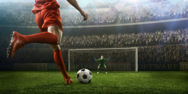 A male soccer player kicks a ball and soccer goalie jumping in motion for a ball while defending his gates on wide angle panoramic image of a outdoor soccer stadium or arena full of spectators under a sunny sky. The image has depth of field with the focus on the foreground part of the pitch. With intentional lensflares. Players are wearing unbranded soccer uniform.