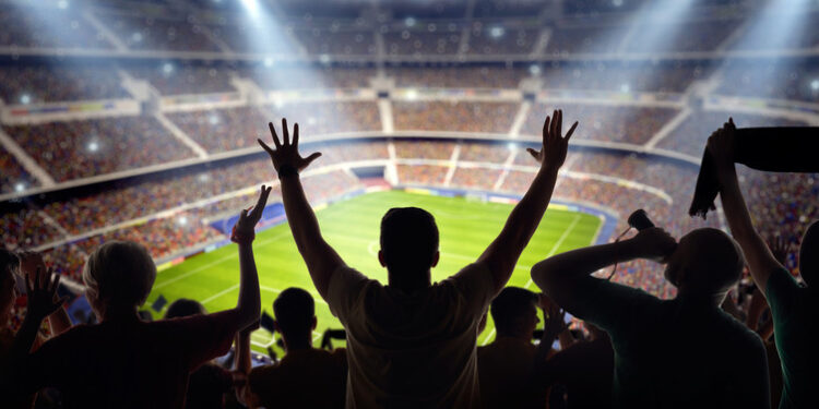 A long-range shot of a stadium field, floodlights and seating. A green field, with painted white lines, is visible in the foreground. On the foreground a group of fans is celebrating a goal. In the background are diffuse out-of-focus stadium seats. Large, bright floodlights are in the top-left and top-right corners of the image.